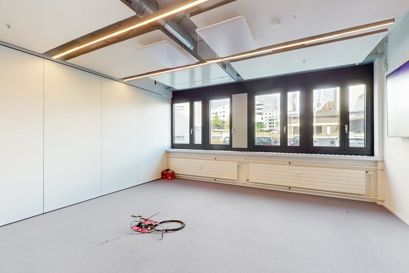 Large open office space with white walls, black framed windows, and a concrete floor. The ceiling has exposed ductwork and lighting fixtures. There is a small red object, possibly a tool or equipment, on the floor.
