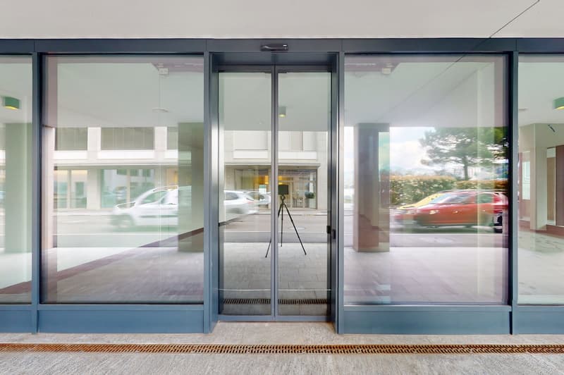 The image shows the entrance of a commercial building with large glass windows and doors. The building has a modern, minimalist design with a gray and white color scheme. The entrance is equipped with automatic sliding doors, and there is a tripod stand i