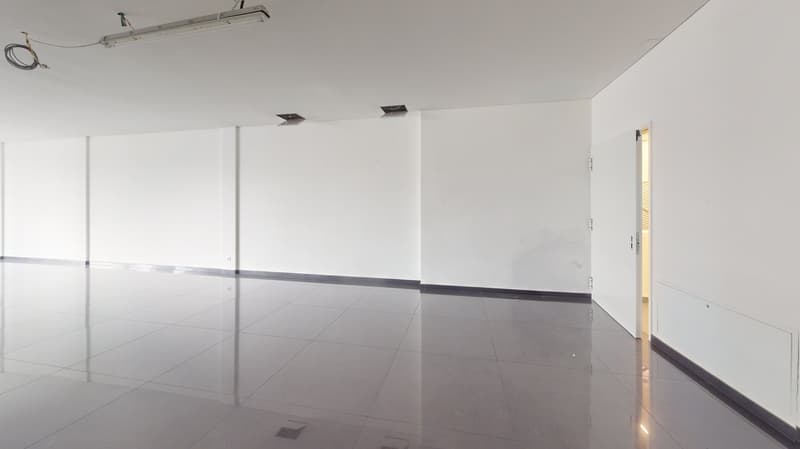 Large open office space with white walls, gray tile flooring, and ceiling-mounted lighting fixtures. The room appears to be empty and ready for use.