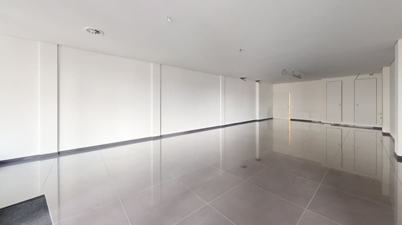 Large open space with white walls, gray tile flooring, and recessed lighting. The space appears to be a commercial or office area, with no visible furniture or fixtures.