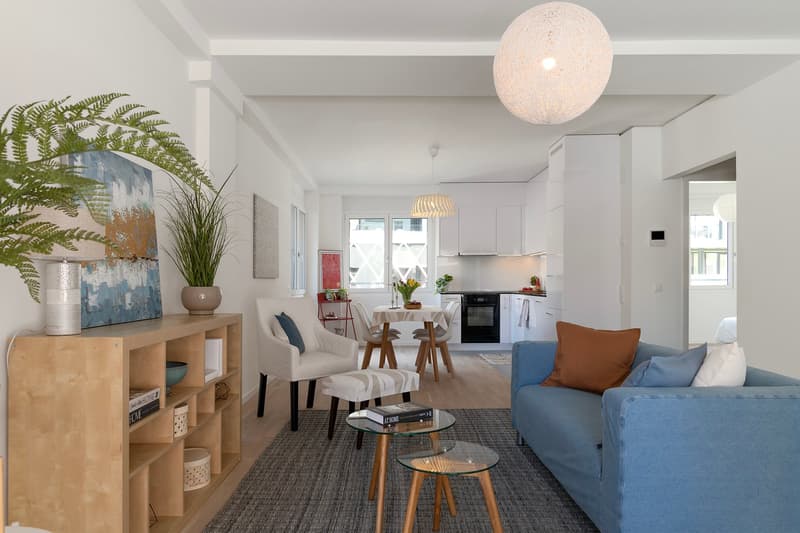 modern living room with blue couch, wooden coffee tables, white chairs, wooden shelves, plants, and a kitchen area