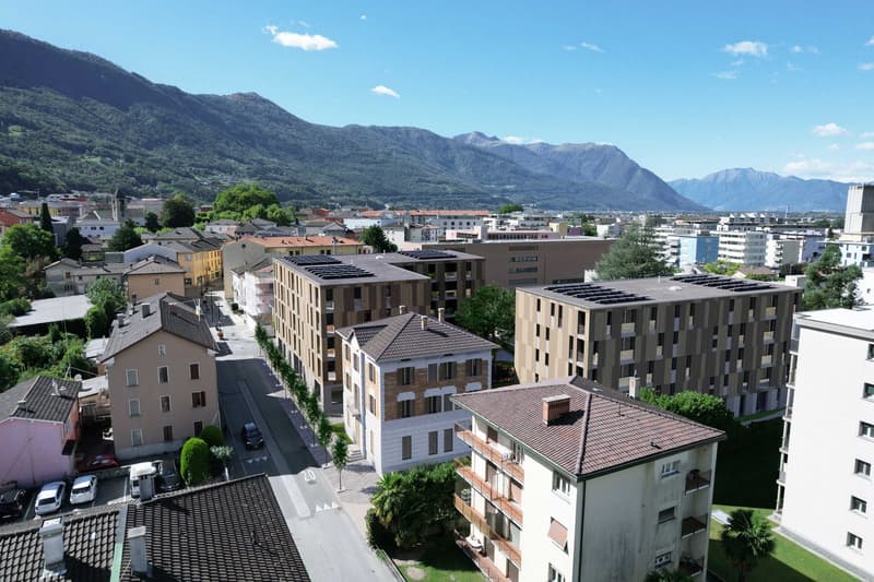 Aerial view of a town with several buildings, mountains in the background, solar panels on some buildings, cars parked on the street, trees and greenery