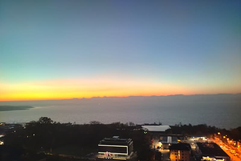 Sunset over the sea with mountain range on the horizon and a city skyline in the foreground.