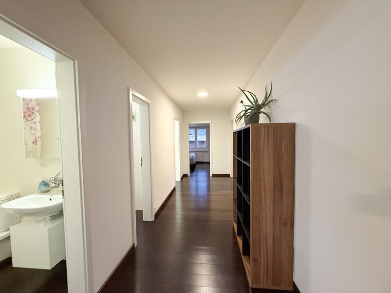 Spacious corridor, tiled floor, white walls, bathroom on the left, a shelf with a plant on the right, a glimpse of a bedroom at the end of the hallway