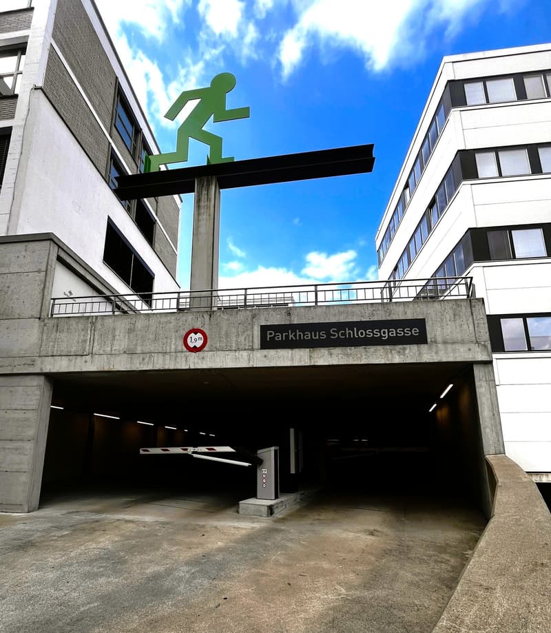A large concrete parking garage with a glass roof, a metal railing on the top, and a pedestrian symbol on a concrete pillar