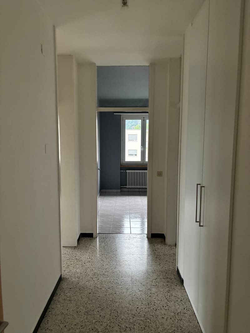 Empty corridor with white walls, tiled floor, and a window with a view of a building.