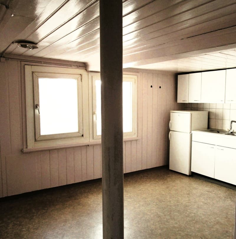 A room with wooden walls and white cabinets and a small refrigerator. The floor is covered with gray tiles. The room has two windows with a white border and an open window in the middle.