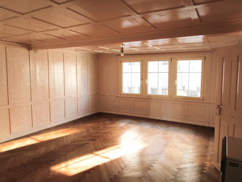 Empty room with wooden floors, walls, three large windows, and a ceiling lamp.