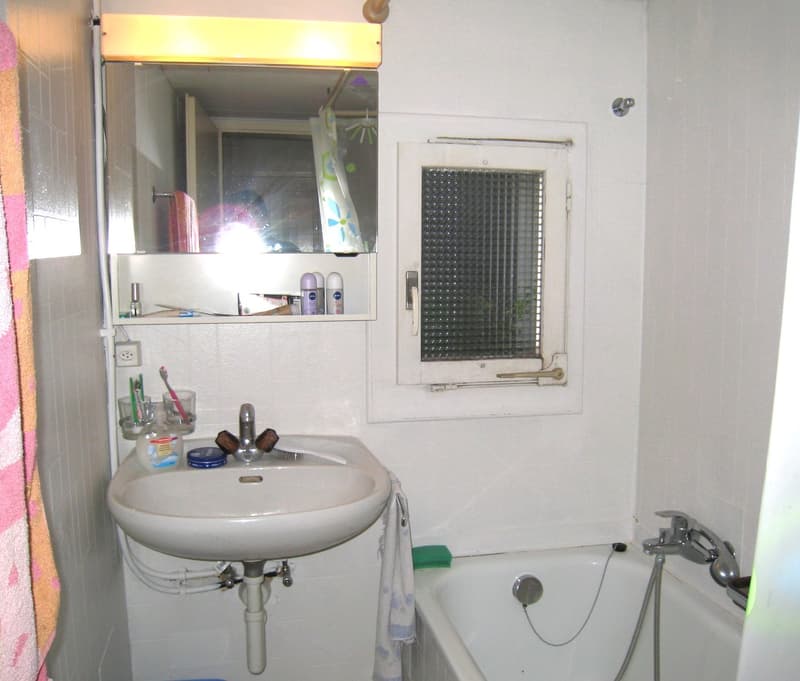 Bathroom with white tiles, a bathtub, a sink, a mirror with a shelf, a window with a grill, and a shower head.