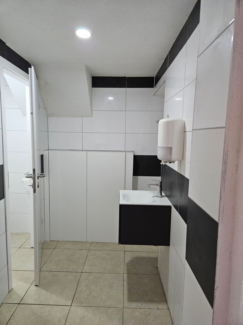 The image shows a modern bathroom with white and black tiled walls, a vanity unit with a sink, and a mirror cabinet above it. The floor is covered with beige tiles. The bathroom appears to be well-lit with recessed lighting in the ceiling.
