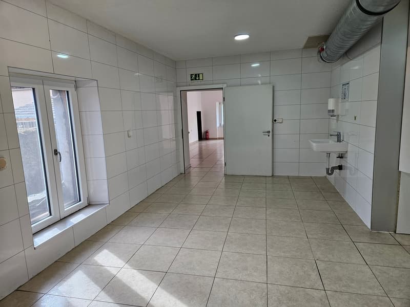 Large open space with white tiled floors, white walls, and a window. The room has a sink and a door leading to another room.