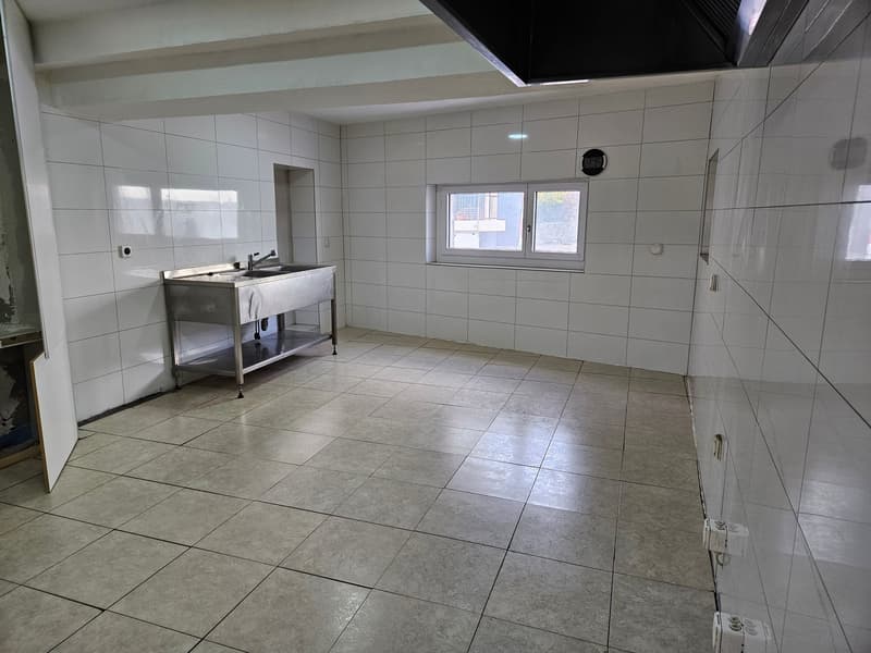 Large open kitchen space with white tiled walls and gray tiled flooring. The room features a stainless steel sink and counter area.