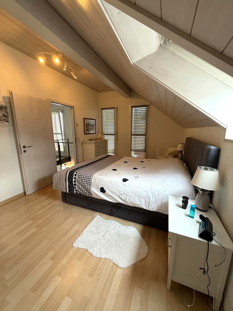 Bedroom with wooden floor, white walls, large bed, nightstand, window with blinds, attic ceiling, skylight, framed picture, rug, wooden cabinet, telephone, lamp