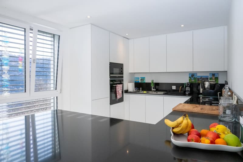 modern kitchen with white cabinets, black countertops, large window, built-in oven and microwave, sink, faucet, chopping board, fruits on the counter