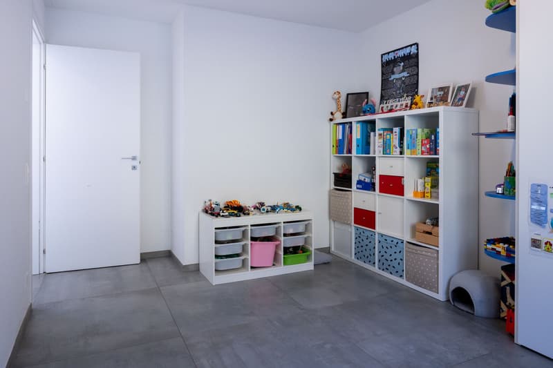Empty room with white walls, gray floor, white door, white shelves, storage boxes, toys, books, and pictures on the shelves.