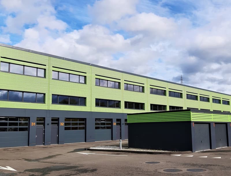 Large green and grey industrial building, several large windows, roll up garage doors on the ground floor, three garages on the side