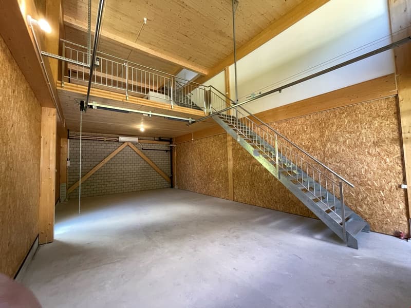 The image shows a spacious garage or storage area with a wooden ceiling, concrete floor, and a metal staircase leading to an upper level. The space appears to be unfinished, with exposed wooden beams and OSB board walls.