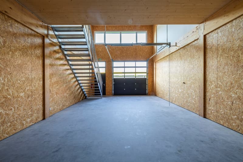 Large open space with concrete floor, wooden walls, and a metal staircase. The space appears to be a garage or workshop area with a garage door visible at the back.