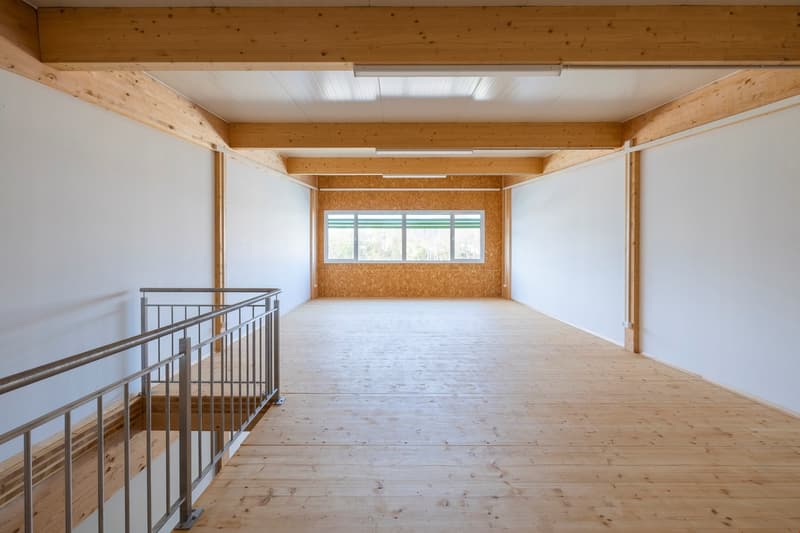 Large open space with wooden beams and ceiling, white walls, and hardwood flooring. The room has a loft-like feel with a metal railing overlooking the main floor area. The room appears to be unfinished or in the process of renovation.