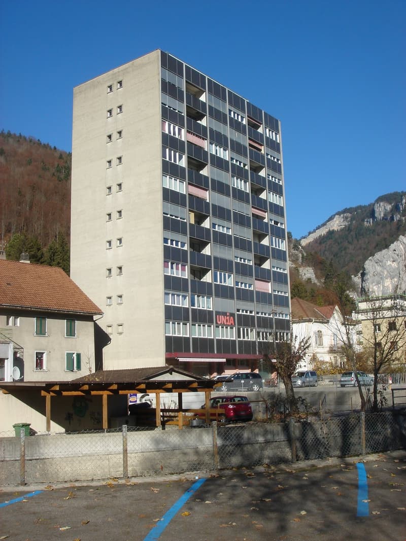 apartment building with 12 floors, windows, balcony on each floor, UNiA sign on the wall, mountains in the background, cars parked in front