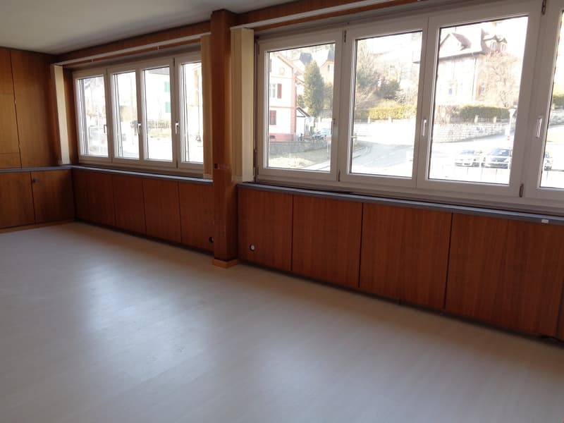 Empty room with white floor, wooden panels, large windows, wooden cabinet with doors