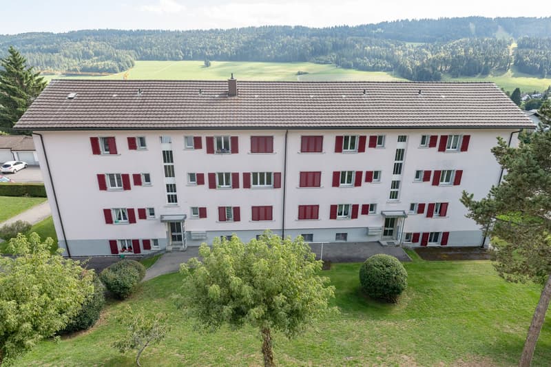 4-story apartment building with red shutters, surrounded by trees and greenery, with a parking area visible in the foreground