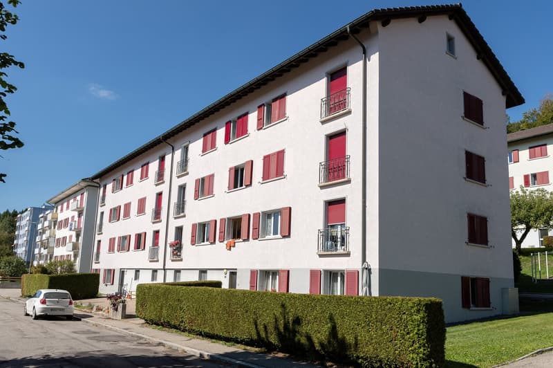 Multi-story apartment building with white exterior walls and red shutters on the windows. The building has balconies on each floor and is surrounded by landscaping with hedges.