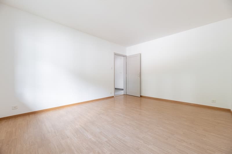 Spacious bedroom with white walls, hardwood flooring, and an open doorway leading to another room.