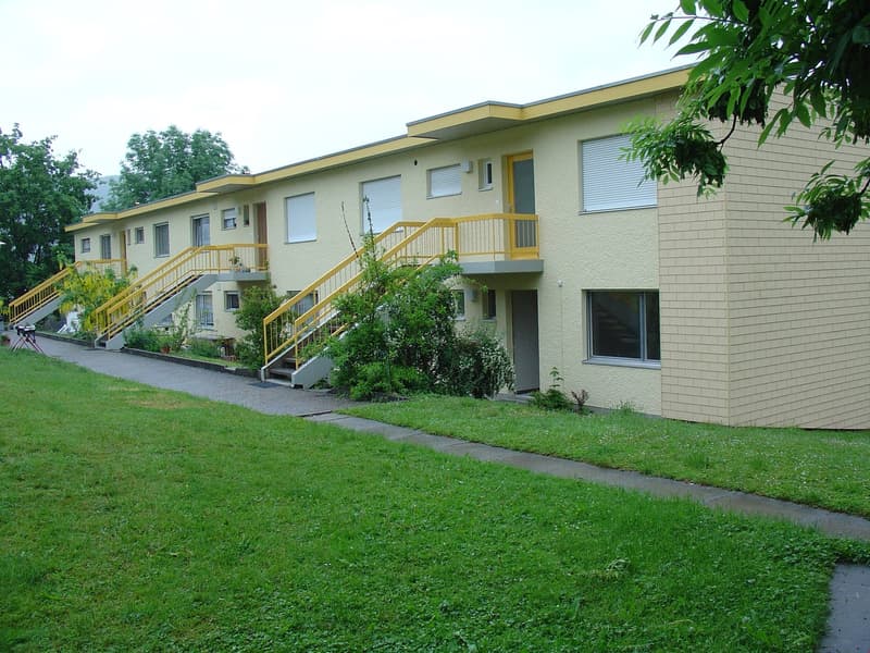 Multi-story apartment building with yellow exterior, balconies, and stairs leading up to the entrances. The building is surrounded by a grassy lawn and trees.