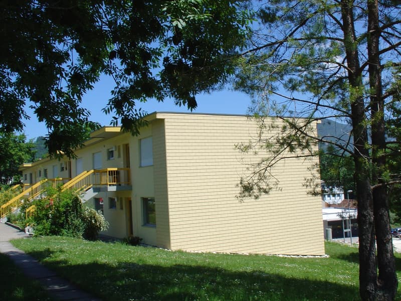 Multi-story apartment building with yellow siding, balconies, and surrounded by trees and greenery