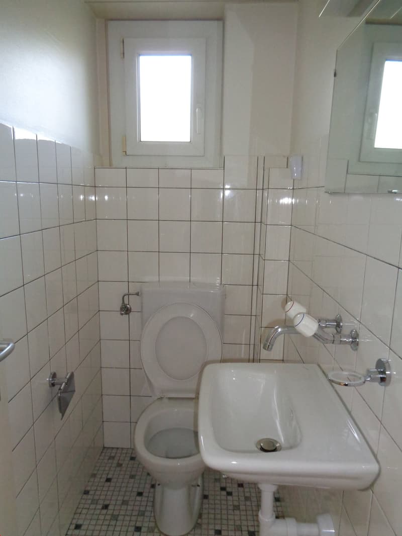 The image shows a bathroom with white tile walls and floor, a toilet, a sink, and a small window.
