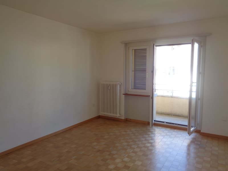 Spacious bedroom with white walls, hardwood flooring, and a sliding glass door leading to a balcony or terrace. The room has a radiator and appears to be in good condition.