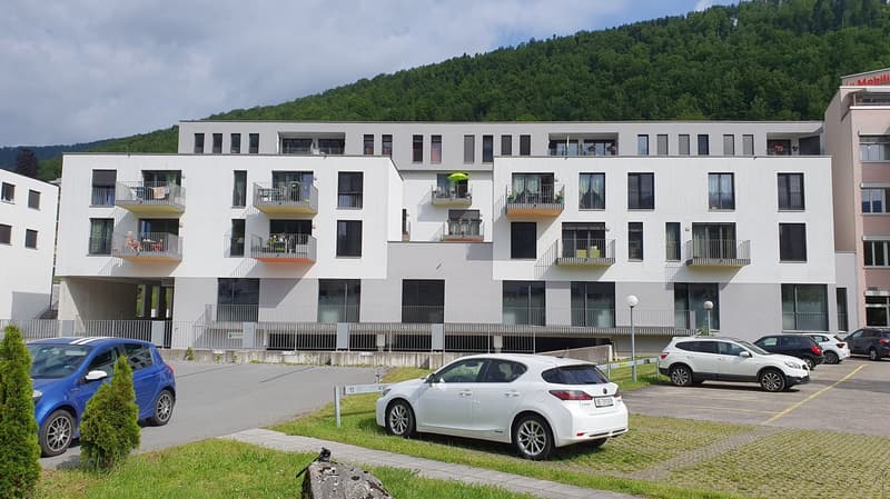 Multi-story apartment building with white exterior, balconies, and parking spaces in front