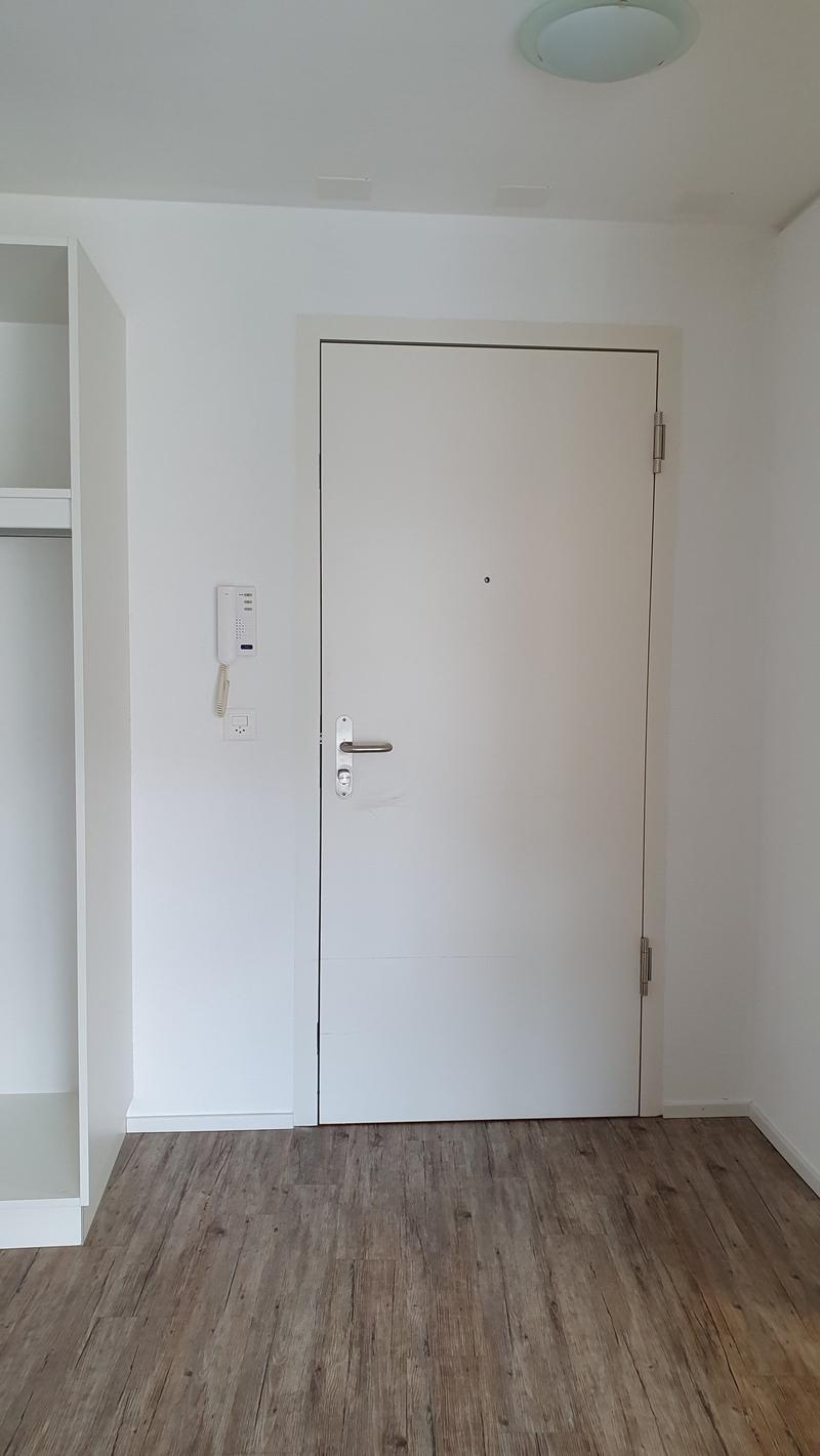 The image shows a simple, minimalist room with white walls and a wooden floor. There is a white door in the center of the frame, with a security intercom system visible next to it. The room appears to be empty, with no furniture or decor visible.
