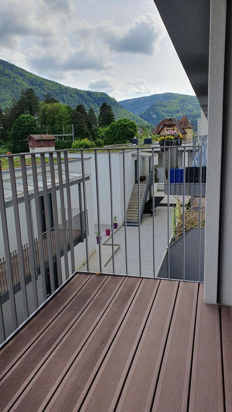 Wooden deck balcony with metal railings, overlooking a mountainous landscape with trees and buildings in the distance.
