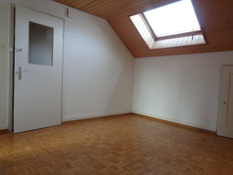 Spacious attic room with wooden ceiling, large window, and hardwood flooring. The room appears to be empty, with a white door and a decorative glass panel on the wall.