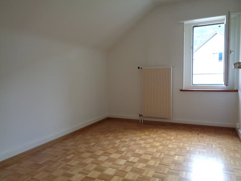 Spacious bedroom with hardwood parquet flooring, white walls, and a radiator. The room has a large window providing natural light.
