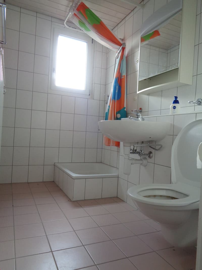 Tiled bathroom with a sink, toilet, and a window. The bathroom has a simple, functional design with white tiles covering the walls and floor.