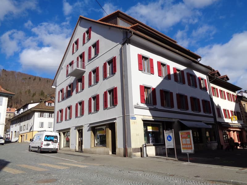 White building with multiple windows, red shutters, balconies, a few shops on the ground floor, cobblestone street