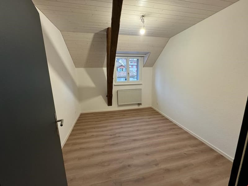 empty room, wooden floors, wooden beams, white walls, ceiling with ceiling light, small window, radiator