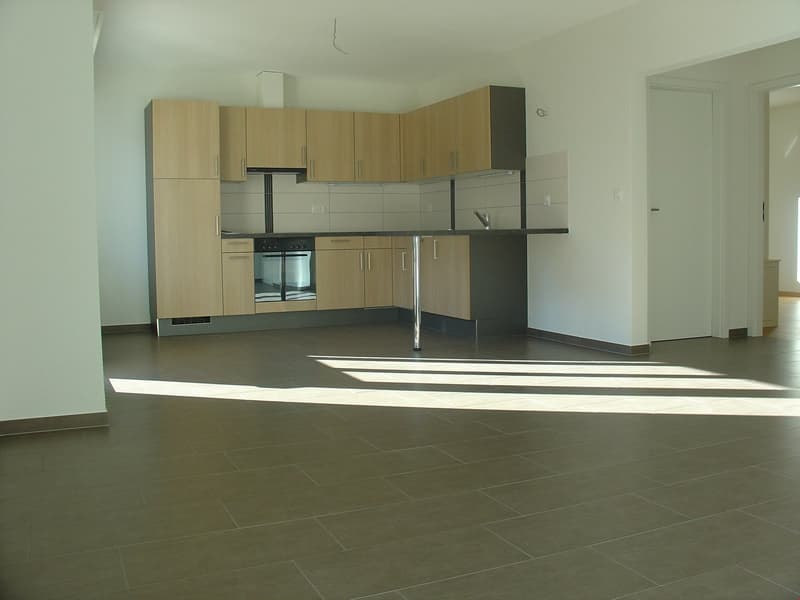 Modern open-concept kitchen with wooden cabinetry, stainless steel appliances, tiled flooring, white walls, kitchen island with sink and faucet, ample counter space, and a sleek stove and oven.