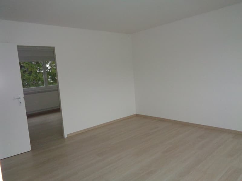 Empty room with white walls, wooden floor, window with blinds, and an open door leading to another room.