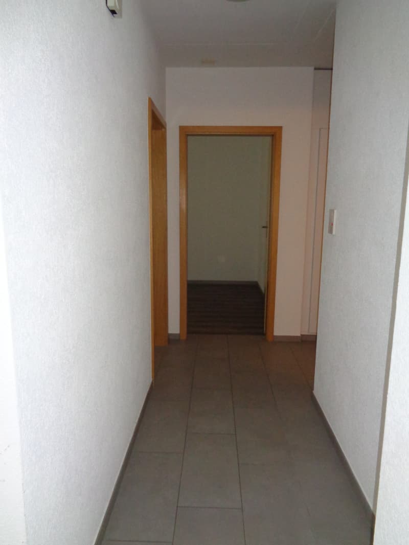 Empty hallway with tiled floor, wooden doors, white walls, and a light on the ceiling.