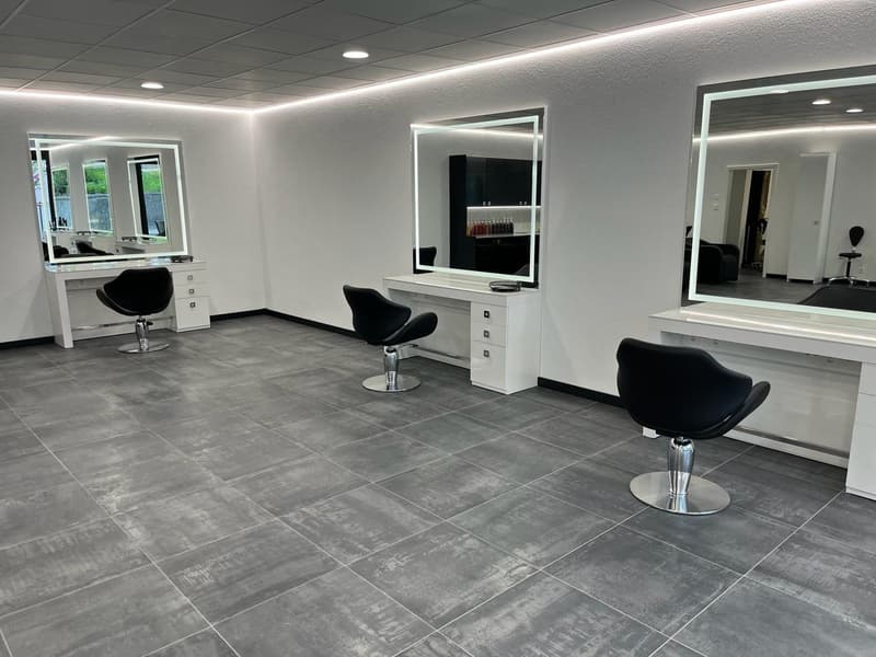 Empty salon with three black swivel chairs, three white desks, three large mirrors, and gray tiled floor.