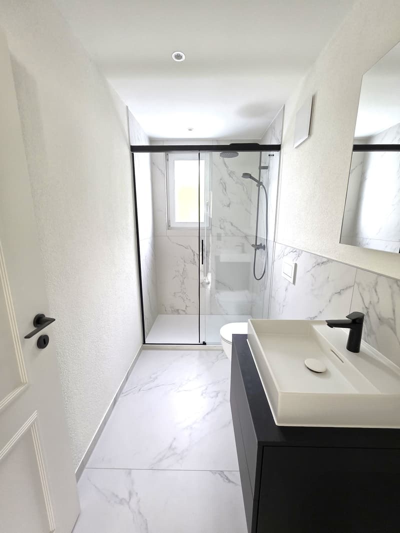 Modern bathroom with marble-like tiles, glass shower enclosure, black fixtures, and a floating vanity with a white countertop