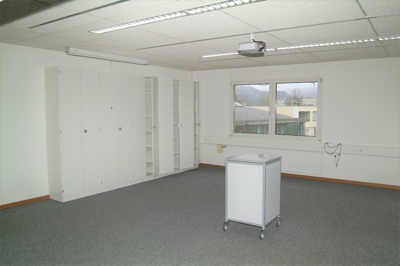 The image shows a spacious office or work space with white walls, a tiled ceiling with recessed lighting, and large windows providing natural light. The room has several built-in storage cabinets or closets along the walls. There is also a small mobile ca