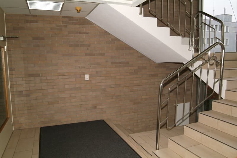 The image shows a staircase with a brick wall on one side and a metal railing on the other. The stairs appear to be made of tile or a similar material. There is a light fixture visible on the ceiling above the stairs.