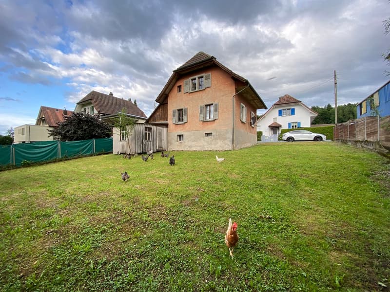 Two-story house with a tiled roof, surrounded by a grassy yard with chickens. There is a garage and a parked car visible on the property.
