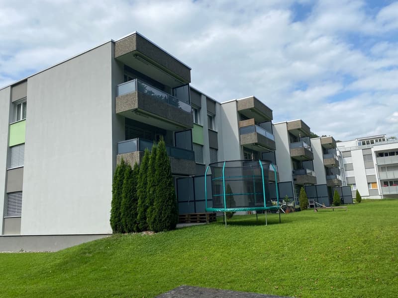Modern apartment building with balconies, concrete exterior, multiple floors, green lawn, and a trampoline