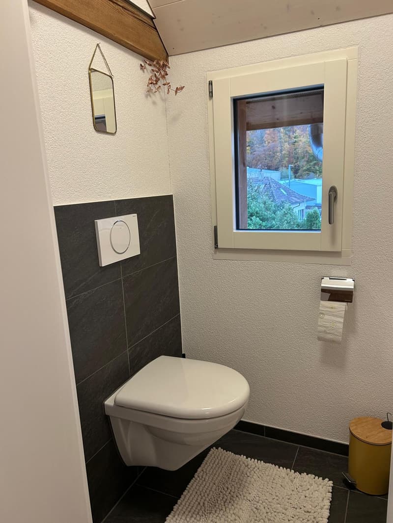 modern bathroom with white toilet, black tiled walls, small window, mirror, white floor mat, trash bin